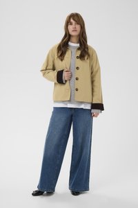 Sieviete stāv, valkājot bēša krāsas pogu jaku ar tumšām aprocēm, pelēku džemperi, baltu kreklu, platām zilām džinsa biksēm un melnus apavus uz vienkārša fona.