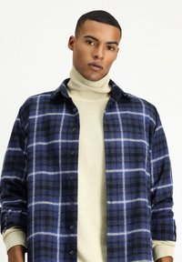 Camicia a quadri blu e nera con chiusura a bottoni, design con colletto e maniche lunghe sopra un dolcevita crema. Tessuto strutturato.