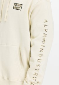 Beige hoodie i mjukt material, med en framficka och en logotyp på lappen. Ärmen har text i camouflagemönster, "ALPHA INDUSTRIES."