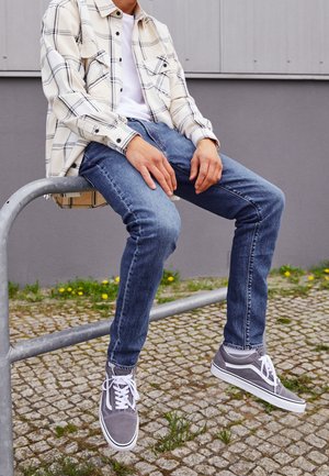 Personne portant des baskets grises, un jean bleu, un t-shirt blanc et une chemise à carreaux crème assise sur une rambarde métallique en extérieur sur un sol pavé.