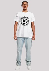 White cotton t-shirt featuring a black printed soccer ball design with "FOOTBALL'S COMING HOME" text, paired with light blue jeans and white sneakers.