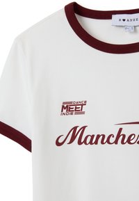 Camiseta de manga corta blanca con cuello y puños burdeos, que presenta el texto burdeos "Dance Meet Indie" y la palabra parcial "Manchester" en la parte frontal.
