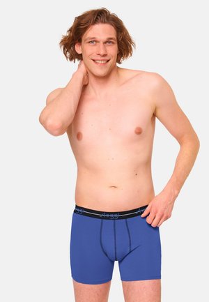 Smiling shirtless man with medium-length hair wearing blue boxer briefs with a black waistband, posing with one hand behind his neck.