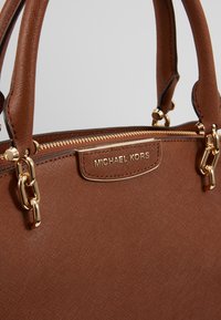 Sac à main en cuir brun texturé avec fermeture éclair dorée, ferrures dorées, doubles poignées et plaque logo "Michael Kors".