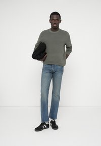 Harmaa teksturoitu villapaita, siniset suorat farkut ja mustat tennarit valkoisilla yksityiskohdilla. Pitää kädessään mustaa teksturoitua esinettä. Yksinkertainen tausta.