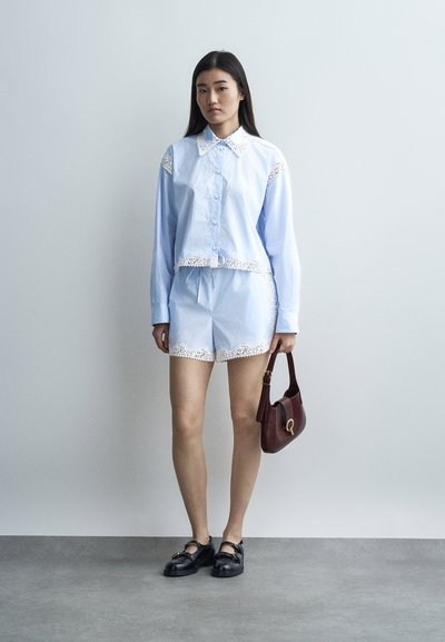 Νεαρή γυναίκα που φοράει ανοιχτό μπλε μακρυμάνικο πουκάμισο και σορτς με λευκή δαντέλα, κρατώντας μια μικρή μπορντό τσάντα, στέκεται πάνω σε γκρι πάτωμα.