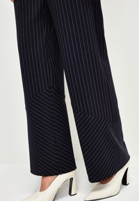 Marineblaue Nadelstreifen-Hosen mit weitem Bein und seitlichen Details, kombiniert mit weißen spitz zulaufenden Pumps mit einem schwarzen Blockabsatz.
