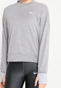 Felpa Nike grigia a maniche lunghe con collo a giro, caratterizzata da un piccolo logo sul petto e polsini texturizzati, abbinata a leggings neri.