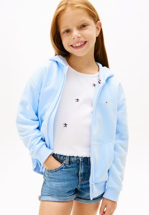 Uśmiechnięta dziewczyna z rudymi włosami, ubrana w jasnoniebieską bluzę na zamek, biały top z małymi wzorami gwiazdek i jeansowe szorty, z rękami w kieszeniach.
