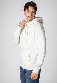 White hooded sweatshirt with a front pocket, soft texture, and embossed branding on the chest and sleeve. Features drawstrings for adjustment.