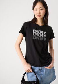T-shirt nera con logo "DKNY" in argento e glitter strutturato. Maniche corte, scollatura rotonda, abbinata a una gonna di jeans blu e una borsa nera.