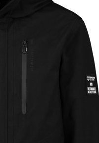 Schwarze Außenjacke mit einer Reißverschlusstasche auf der Brust, strukturiertem Stoff und einem Aufnäher mit der Aufschrift "ULTIMATIVE ABLEHNUNG" sowie Logodetails.
