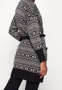 Pletený dlouhý kardigan v námořnické modři a krémové barvě, zdobený geometrickými vzory, s páskem v pase a bočními kapsami. Žebrované texturované manžety a lem.