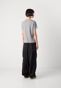 En person med kort hår står med ryggen til iført en grå t-skjorte, løse svarte cargo-bukser og brune joggesko mot en ensfarget bakgrunn.