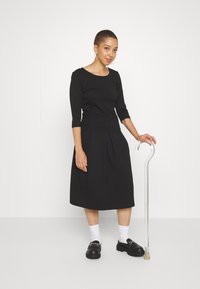 Femme aux cheveux courts portant une robe noire, des chaussettes blanches et des chaussures noires, debout tenant une canne argentée à poignée courbée.