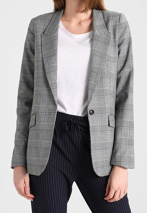 Person iført en grå ternet blazer over en hvid skjorte med sorte bukser med smalle striber, bundet i livet.