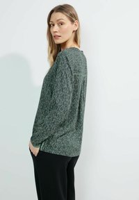 Blusa verde con un motivo strutturato, maniche lunghe e un fit rilassato. Presenta una discreta piega sul retro e un orlo arrotondato. Abbinata a pantaloni neri.