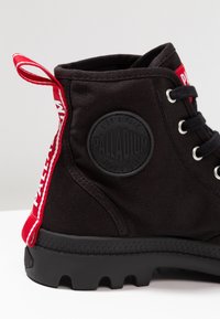 Bottine en toile noire avec semelle en caoutchouc, ornée d'un logo embossé rond, d'une languette rouge et de lacets noirs. Accent latéral en rouge et blanc.