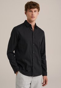 Črna majica z dolgimi rokavi v teksturirani tkanini, z gumbom na sprednji strani in klasičnim ovratnikom. Nosijo jo z svetlo sivo vzorčasto hlačami.