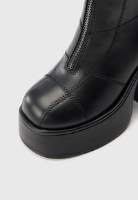 Bota de cano curto em pele preta com bocal arredondado, com costura contrastante e fecho de correr lateral. Sola tipo plataforma grossa para maior altura.