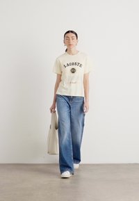 Krem majica s logotipom "LACOSTE", uparen sa plavim širokim trapericama i bijelim tenisicama. Model nosi bež torbu.