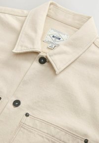 Crèmekleurige denimjack met een kraag, twee voorzakken, zwarte drukknoopjes en een merklabel binnenin de kraag.