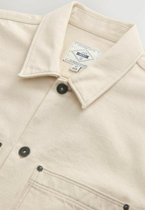 Cremfarbene Jeansjacke mit kragenförmigem Ausschnitt, zwei vorderen Taschen, schwarzen Druckknöpfen und einem Markenetikett im Kragen.