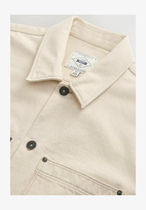 Giacca di denim color crema con colletto, due tasche frontali, bottoni neri a pressione e un'etichetta con marchio all'interno del colletto.