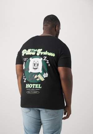 Mężczyzna w czarnej koszulce z kreskówkową postacią poduszki i tekstem o "The Pillow Fortress Hotel" promującym sen.