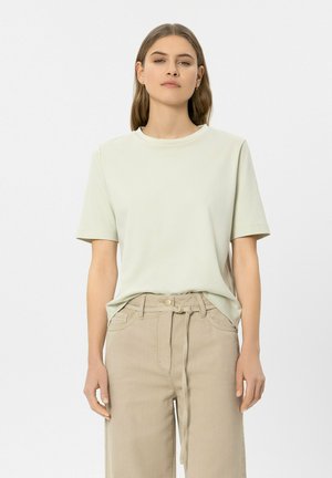 Junge Frau mit glatten braunen Haaren trägt ein hellbeiges kurzärmeliges Hemd, das in eine hochtaillierte beige Hose mit Stoffgürtel gesteckt ist.