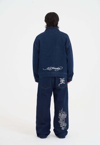 Džínová bunda a kalhoty v tmavě modré barvě. Obsahují bílé vyšívané logo a podrobný design draka na nohavici kalhot. Volný střih, klasický design.