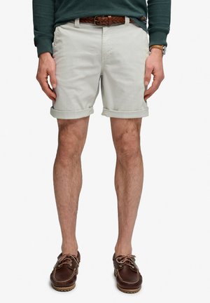 Man met beige opgerolde shorts, bruine gevlochten riem, groen overhemd met lange mouwen, bruine leren bootschoenen en een horloge.