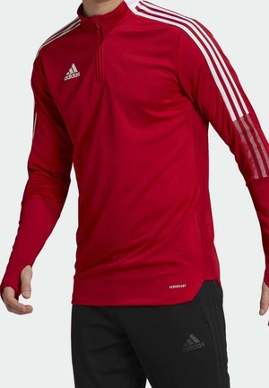 Mann trägt rotes Adidas Langarmshirt mit weißen Streifen und schwarze Adidas-Hose, steht mit leicht ausgestreckten Armen.