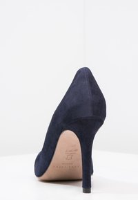 Marinblå mocka högklackad sko med spetsig tå och en smal stilettklack. Slät yta och minimalistisk design, med en logotyp inuti.