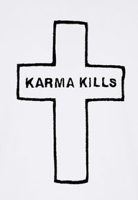 Tissu blanc avec une croix noire brodée et les mots "KARMA KILLS" en lettres épaisses. La texture semble en relief et floue.
