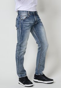 Jeans de denim azul con un corte ajustado, ligero desgaste, sutileza en los pliegues, cinco bolsillos y costuras contrastantes. Combinados con zapatillas negras.