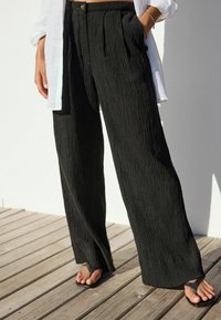 Pantaloni negri cu tăieturi late dintr-un material texturat, cu pliuri frontale și un talie cu nasture, asortați cu sandale negre de tip flip-flop.