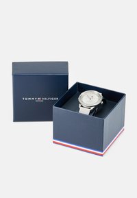 Tommy Hilfiger laikrodis tamsiai mėlyname dėkle su sidabriniu skydeliu, baltu dirželiu ir logotipo detale. Dėklas turi raudonos, baltos ir mėlynos spalvų juostų akcentą.