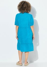 Türkises Leinenkleid mit kurzen Puffärmeln, gestuftem Saum und lockerer Passform. Getragen mit flachen, gewebten Slipper-Sandalen.