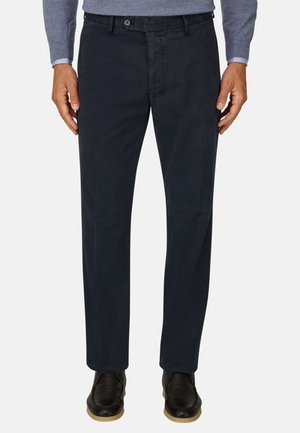 Homme portant un pantalon droit bleu marine foncé, des chaussures noires à semelles claires et un pull gris avec les poignets de chemise visibles.
