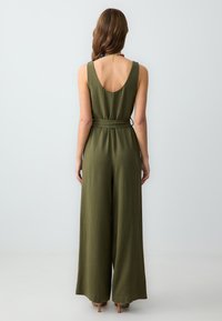 Combinaison vert olive avec un dos dénudé, une coupe ample et un détail de taille nouée. Texture lisse avec un tissu léger.