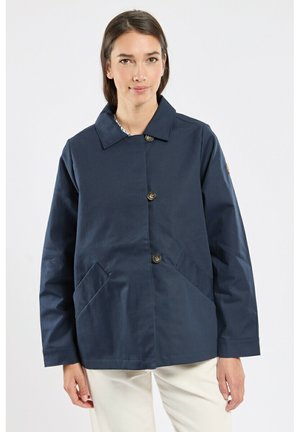 Femme portant une veste bleue marine boutonnée avec un col et des poches avant inclinées, assortie à un pantalon beige clair sur un fond blanc.