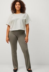 Krullendharige vrouw in een losse beige t-shirt, olijfgroene strakke broek en zwarte schoenen staat tegen een eenvoudige lichte achtergrond.