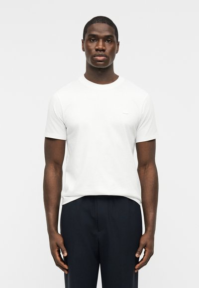 Μαύρος άνδρας όρθιος, φορώντας απλό λευκό μπλουζάκι με κοντά μανίκια και σκούρο παντελόνι μπροστά σε απλό ανοιχτό φόντο.