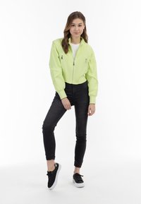 Giubbotto bomber verde neon tagliato con tasche con zip, polsini a coste, indossato sopra una camicia bianca, abbinato a jeans skinny neri e sneakers nere.
