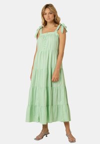 Muntgroene tiered maxi-jurk met verstelbare strikbandjes, vierkante halslijn en knoopdetails op het lijfje. Geweven textuur, vloeiende silhouet.