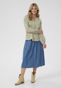 Hellgrüne bestickte Bluse mit langen Ärmeln und Knopfdetails, kombiniert mit einem fließenden blauen Jeansrock und beigen Cowboy-Stiefeln.