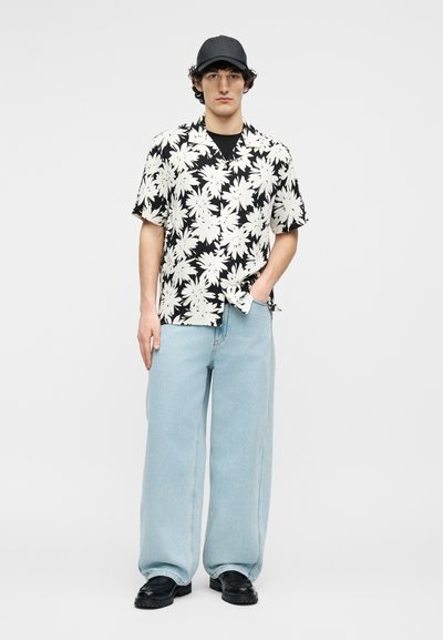 Jeune homme portant une casquette noire, une chemise à manches courtes noire et blanche à motif floral, un jean bleu clair à jambes larges, et des chaussures noires, debout les mains dans les poches.