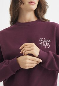 Sudadera burdeos con cuello redondo, puños acanalados y bordado en el pecho izquierdo del texto "Chelsea Peers" en blanco. Textura suave.