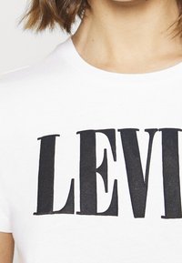 T-shirt en coton blanc avec un grand logo noir "LEVIS" sur la poitrine. Le tissu semble lisse avec un col rond.